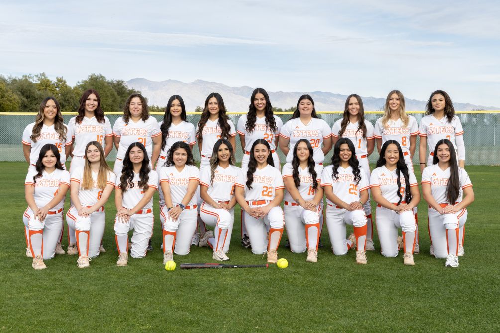 Photo: Team photo of a women&rsquo;s softball team posing on a field in white and orange &ldquo;Aztecs&rdquo; uniforms with mountains visible in the background. Players are arranged in two rows, with one row standing and another kneeling in front. A softball bat and two softballs are placed on the grass in front of the group.
