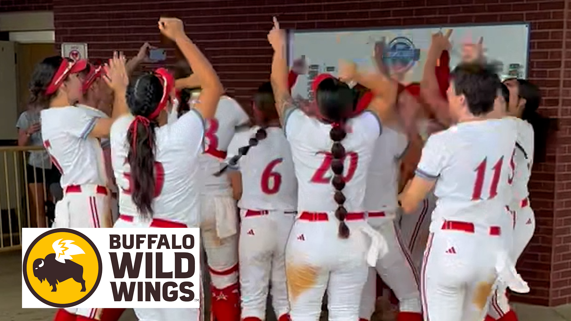 Photo: Softball players in white and red uniforms celebrating together in a tunnel or dugout entrance area with their arms raised in the air. Several players are visible from the back with jersey numbers showing, while teammates gather in a group celebration near a brick wall. A Buffalo Wild Wings logo graphic is displayed in the bottom left corner of the image.
