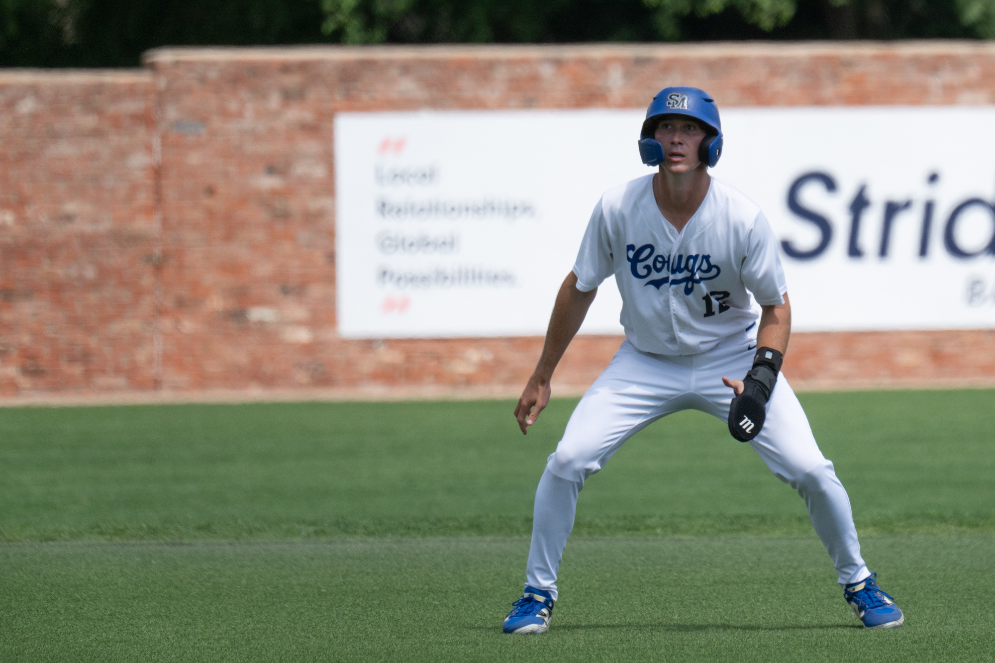 Photo: Baseball player in a white &ldquo;Cougs&rdquo; uniform leading off a base during a game. The athlete is wearing a blue batting helmet and protective sliding mitt while standing in an athletic ready position on the field. A brick wall and blurred advertisement sign are visible in the background.

