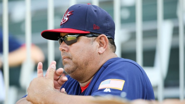 Photo: A baseball coach wearing a navy USA Baseball cap, sunglasses, and a navy pullover with an American flag patch watches the action from the dugout with his hands clasped. The background shows blurred stadium seating and another person out of focus.
